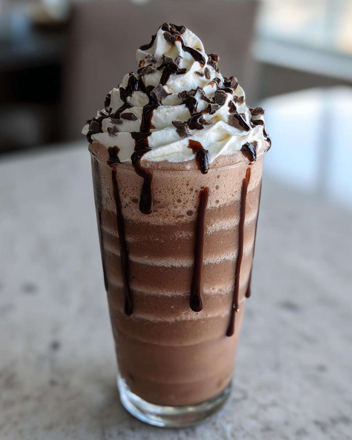 A tall glass of rich frozen hot chocolate topped with whipped cream, chocolate drizzle, and shavings.