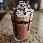 A tall glass filled with blended frozen hot chocolate topped with whipped cream, chocolate syrup, and shavings.