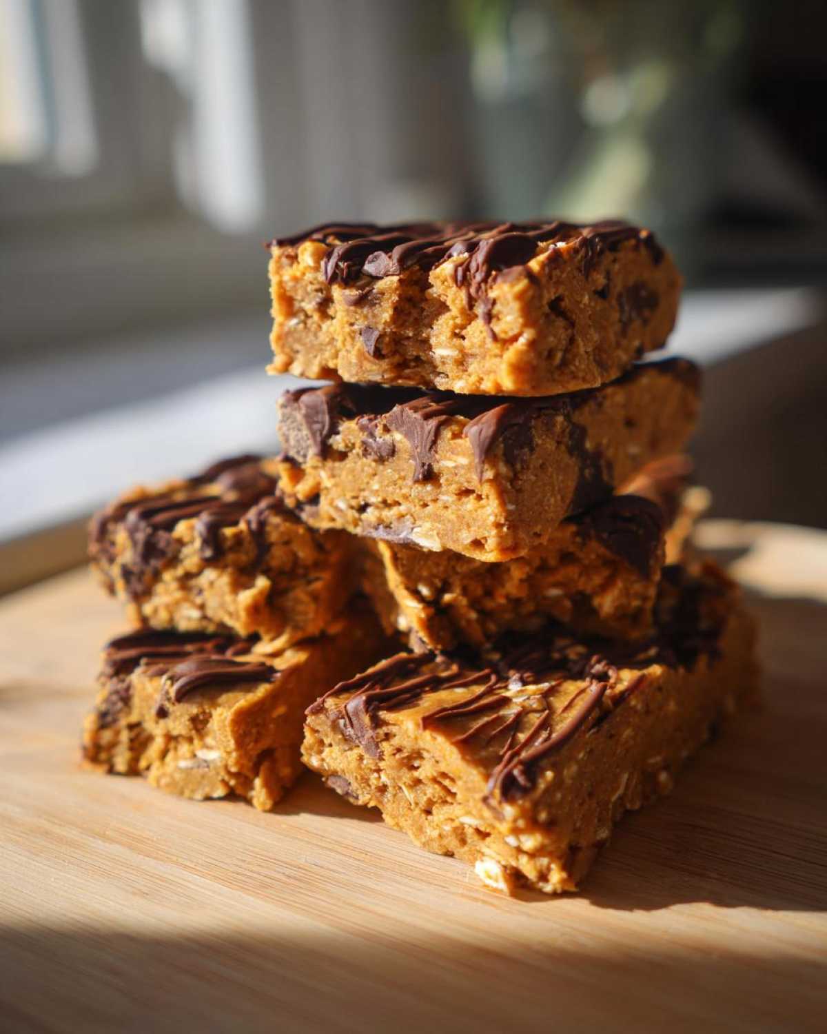 A stack of homemade, peanut butter colored protein bars topped with drizzled dark chocolate, sitting on a wooden board.