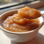 Close-up of homemade applesauce being lifted from a white bowl on a spoon, showing its chunky texture.