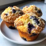 Three golden blueberry muffins topped with a crunchy sugar streusel sitting on a white plate.