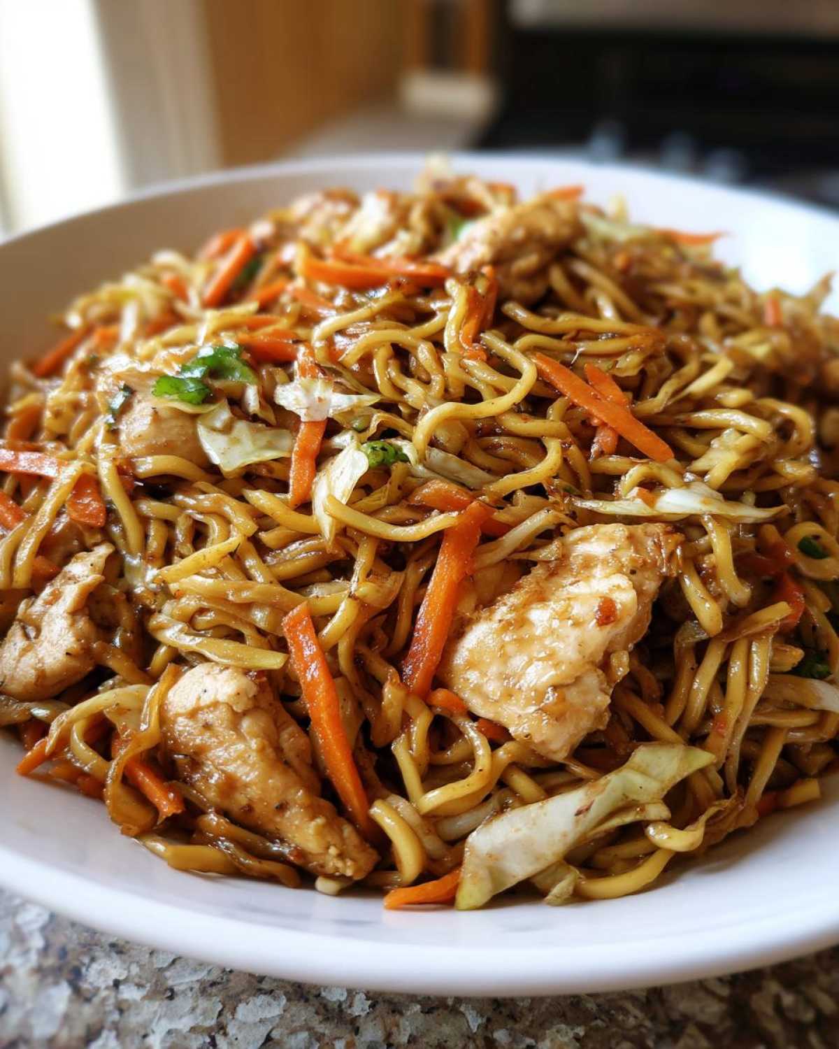 A close-up view of a white bowl filled with amazing chicken chow mein recipe, featuring noodles, chicken pieces, carrots, and cabbage.
