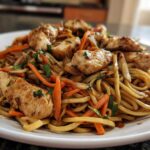 Close-up of a white plate filled with glistening chicken chow mein recipe, featuring thick noodles, sliced chicken breast, and julienned carrots.