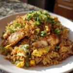 A close-up of a white bowl filled with savory chicken fried rice, featuring pieces of chicken and scrambled egg, topped generously with fresh green onions.