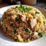 A mound of savory chicken fried rice with visible chunks of chicken, scrambled egg, and green onions on a white plate.