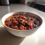 A close-up of a steaming white bowl filled with rich, hearty crockpot chili, featuring ground beef and kidney beans.