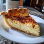 A close-up of a thick slice of creamy egg custard pie dusted with cinnamon on a white plate.