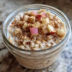 A glass jar filled with creamy apple cinnamon overnight oats, topped with diced red apples and sprinkled cinnamon.