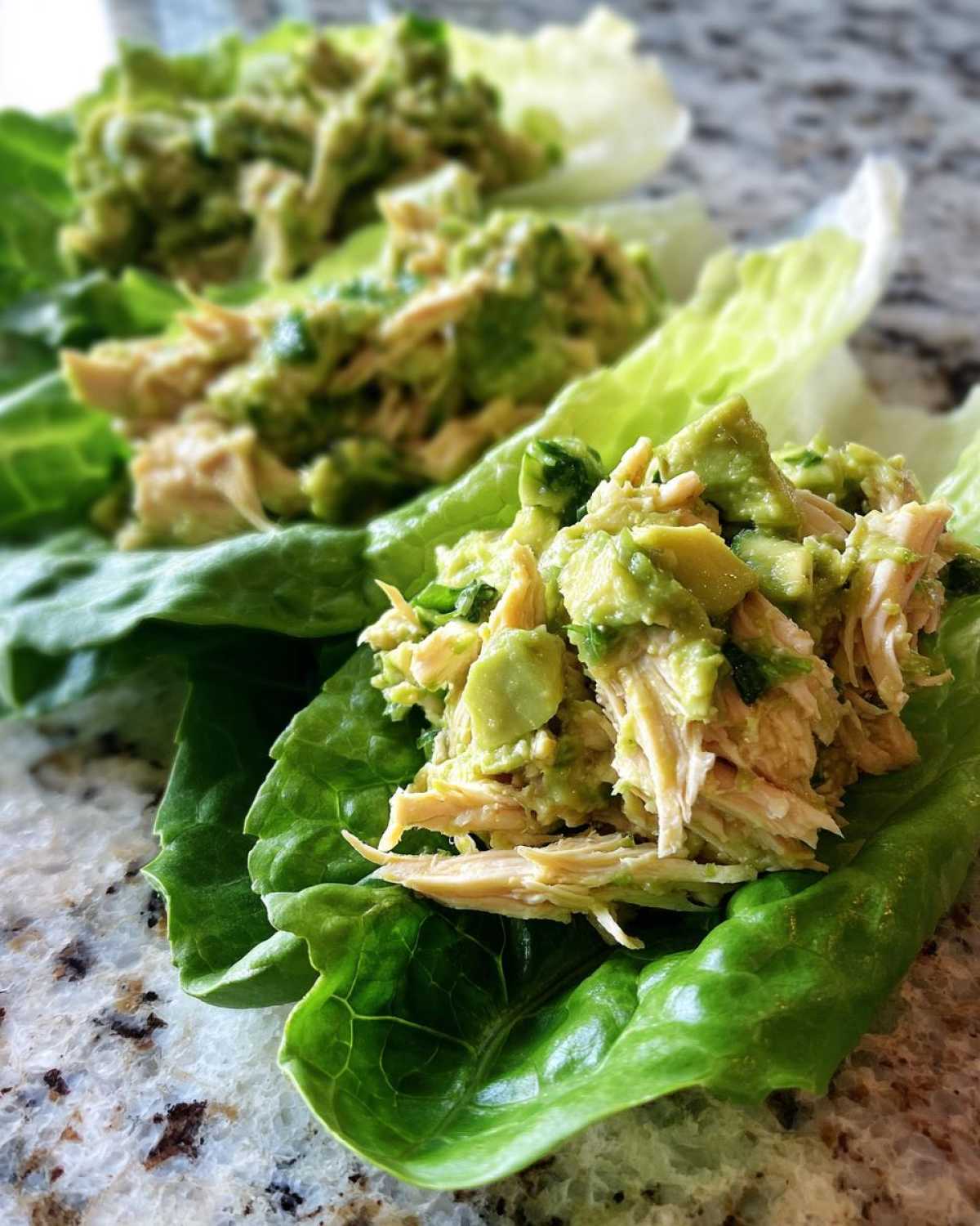 Close-up of creamy avocado chicken salad served in crisp green lettuce cups on a granite counter.