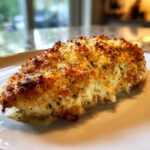 A close-up of a perfectly baked caesar chicken breast with a golden, cheesy, and herbed crust resting on a white plate.