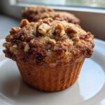 Close-up of a perfectly baked banana nut muffin topped with crunchy walnuts, sitting on a white plate.