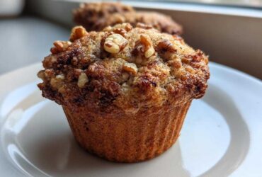 Close-up of a perfectly baked banana nut muffin topped with crunchy walnuts, sitting on a white plate.