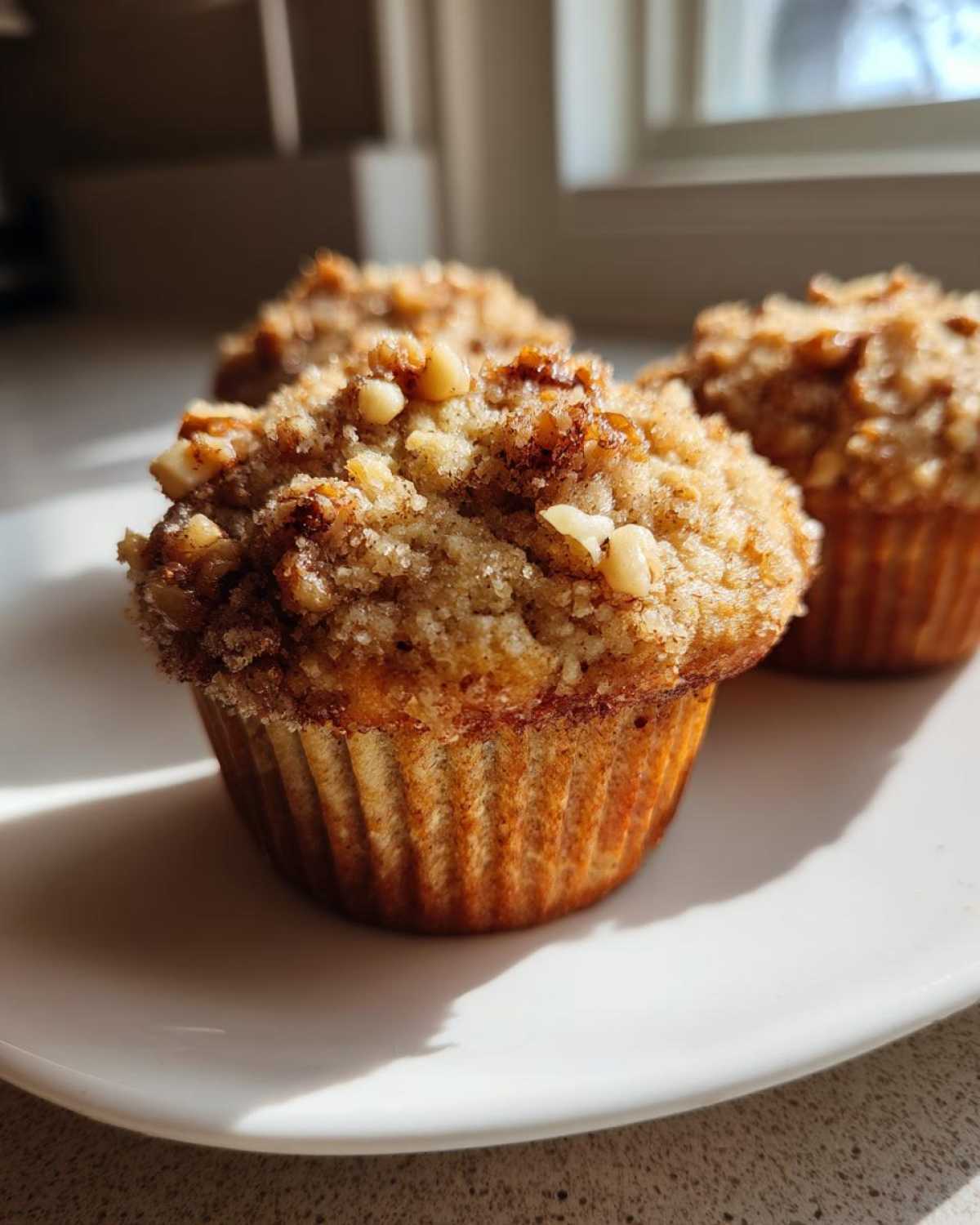 A close-up of freshly baked banana nut muffins with a crunchy, sugary nut topping, sitting on a white plate.