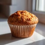 A single, golden-brown banana protein muffin sitting in a white paper liner, highlighted by bright sunlight.