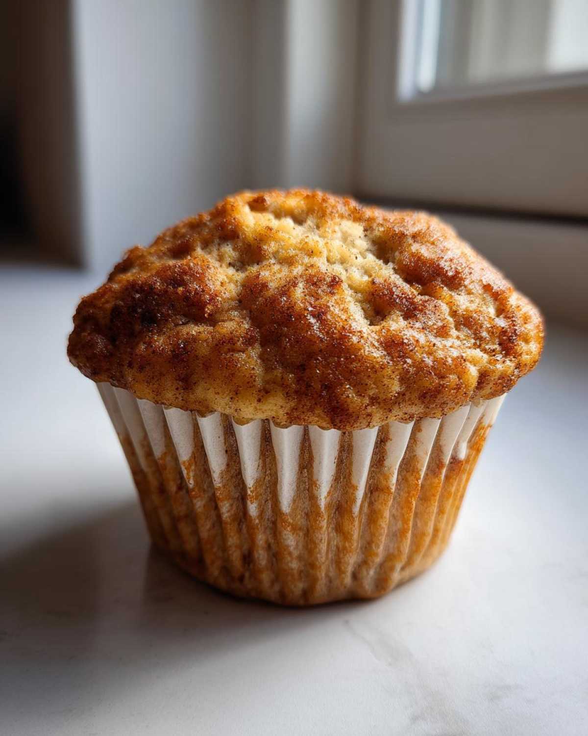 A single, freshly baked banana protein muffin with a golden-brown, cinnamon-dusted top, sitting in a white paper liner.