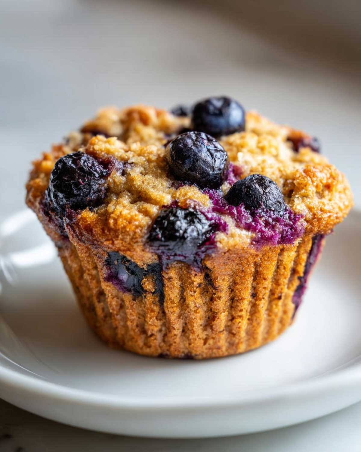 A single, golden brown blueberry protein muffin topped with fresh blueberries, sitting on a white plate.
