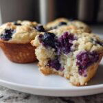 A close-up of a blueberry protein muffin with a bite taken out, revealing the moist interior and purple berries.