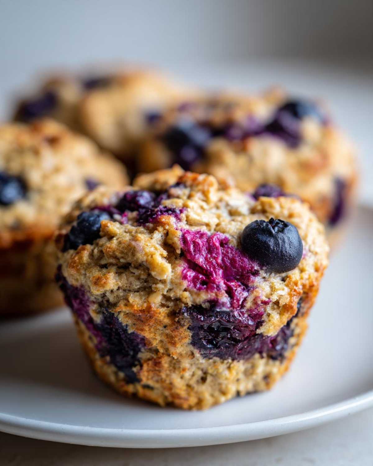 A close-up of a fresh, oat-topped blueberry protein muffins sitting on a white plate.