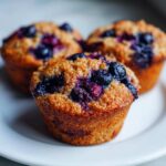 Three freshly baked blueberry protein muffins with golden tops sitting on a white plate.