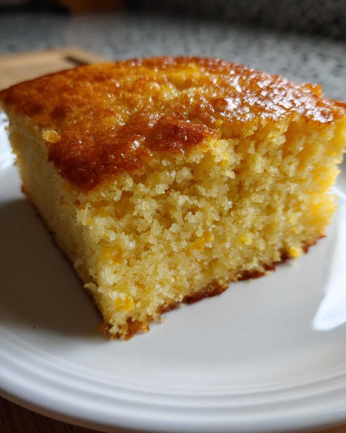 Close-up of a square slice of moist buttermilk cornbread with a shiny, golden-brown crust on a white plate.