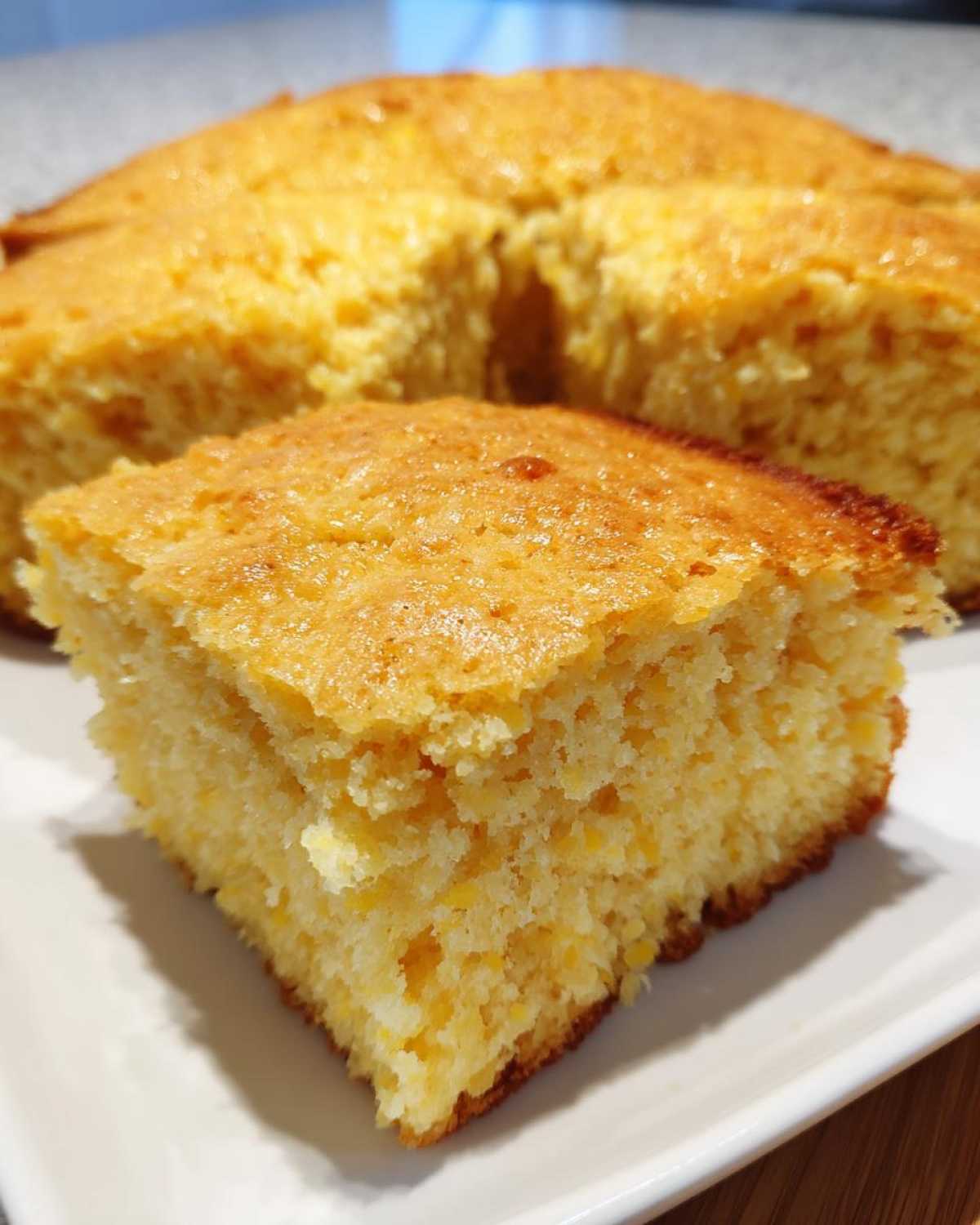 A close-up of a square slice of golden buttermilk cornbread resting on a white plate, with the rest of the round cornbread visible behind it.