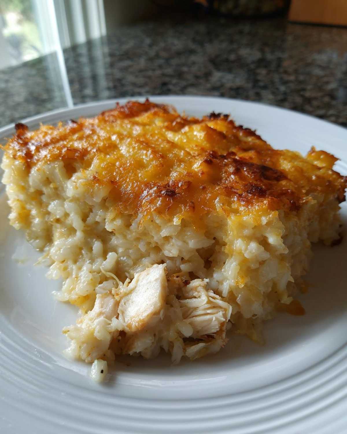 A square serving of cheesy chicken and rice casserole with a golden brown, bubbly cheese topping on a white plate.