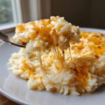 A fork lifts a scoop of creamy, cheesy rice, showing long, satisfying cheese pulls from the melted cheddar topping.