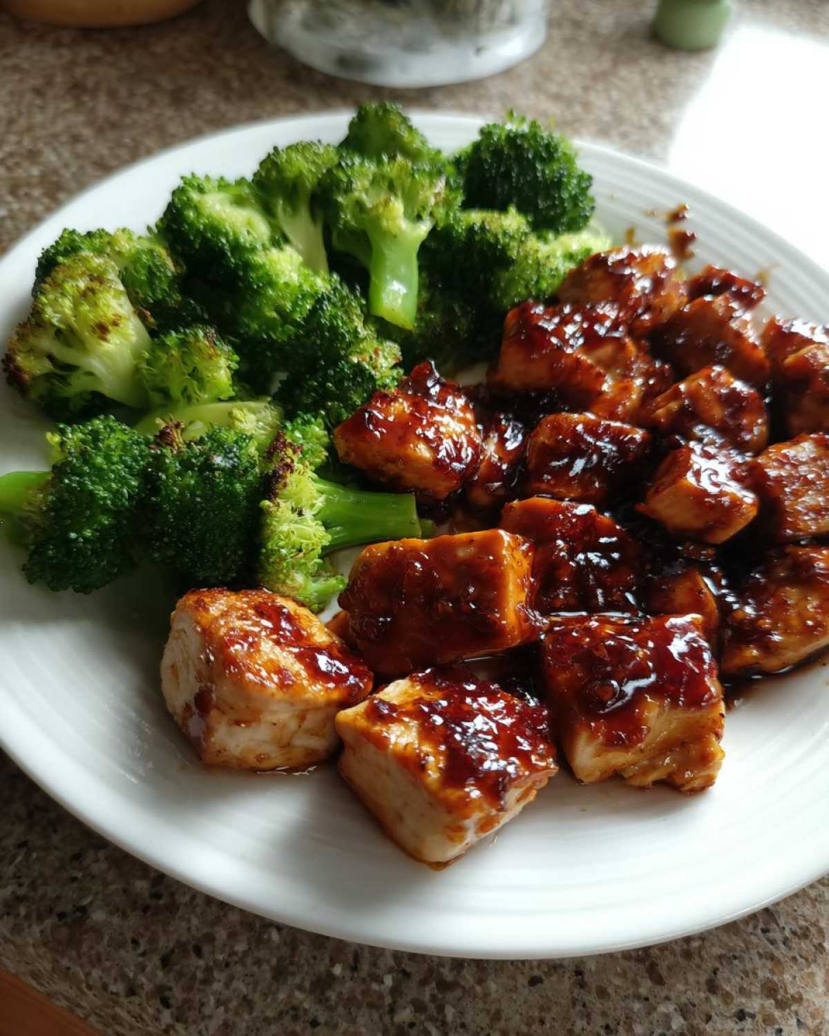 A white plate featuring glazed chicken pieces next to bright green steamed or roasted chicken and broccoli.