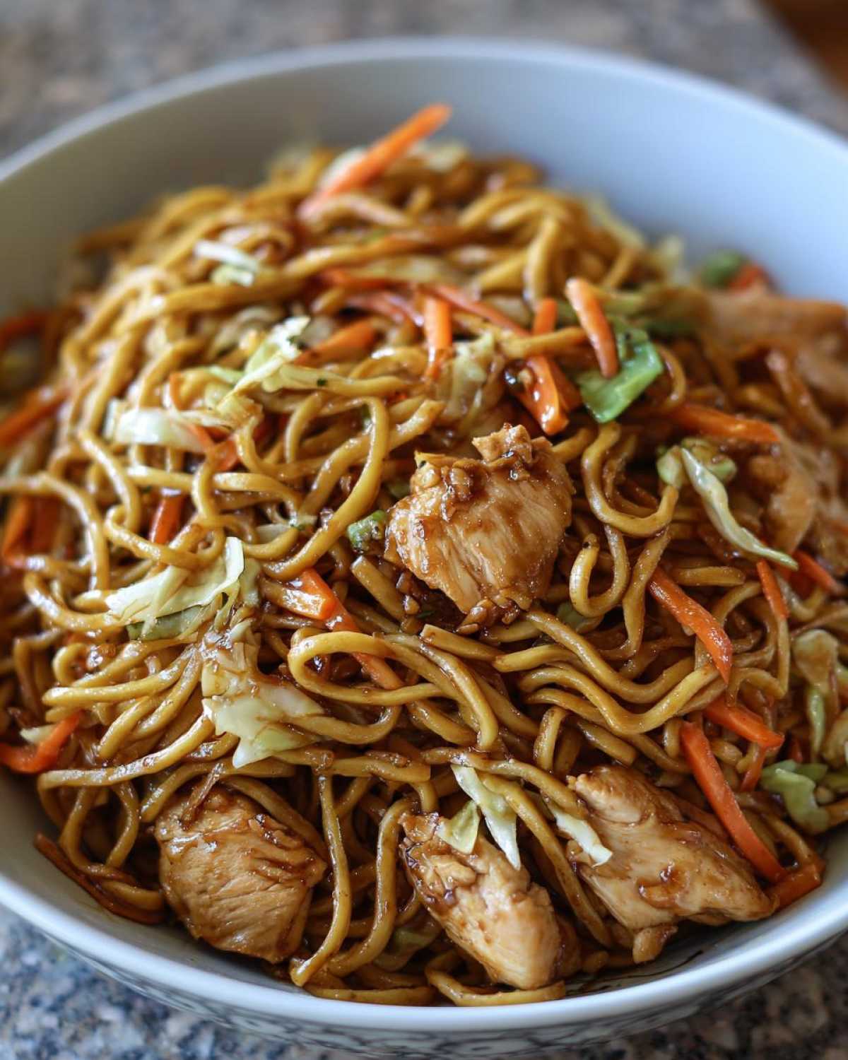 A close-up overhead shot of a bowl filled with glossy noodles, chunks of chicken, and shredded carrots and cabbage from the chicken chow mein recipe.