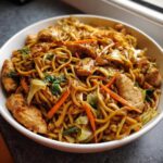 A close-up of a white bowl filled with a savory chicken chow mein recipe, featuring noodles, chicken pieces, carrots, and cabbage.