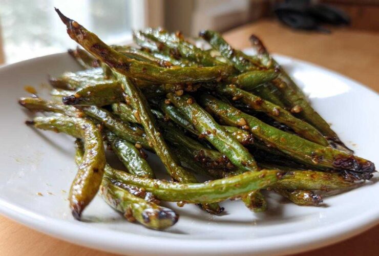 A close-up of blistered, glossy chinese green beans with garlic sauce piled on a white plate.