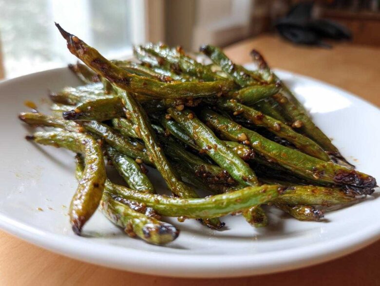 A close-up of blistered, glossy chinese green beans with garlic sauce piled on a white plate.