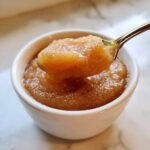 A spoonful of rich, brown homemade applesauce being lifted from a small white bowl.
