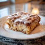 A close-up of a moist square slice of cinnamon roll casserole topped with thick white icing on a white plate.