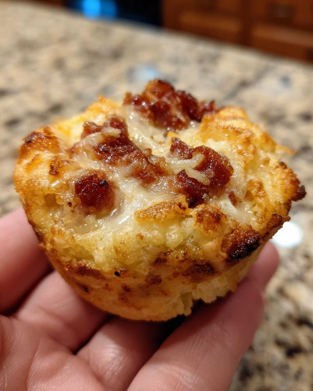 A close-up shot of a freshly baked pizza muffin held in a hand, showing melted cheese and meat toppings.
