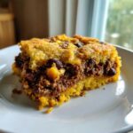 A close-up slice of savory cowboy cornbread casserole showing a thick layer of chili filling between two cornbread layers.