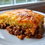A hearty slice of cowboy cornbread casserole with a golden top and savory ground meat filling on a white plate.