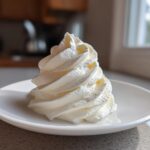 A tall, perfectly piped swirl of white cream cheese frosting sitting on a small white saucer.