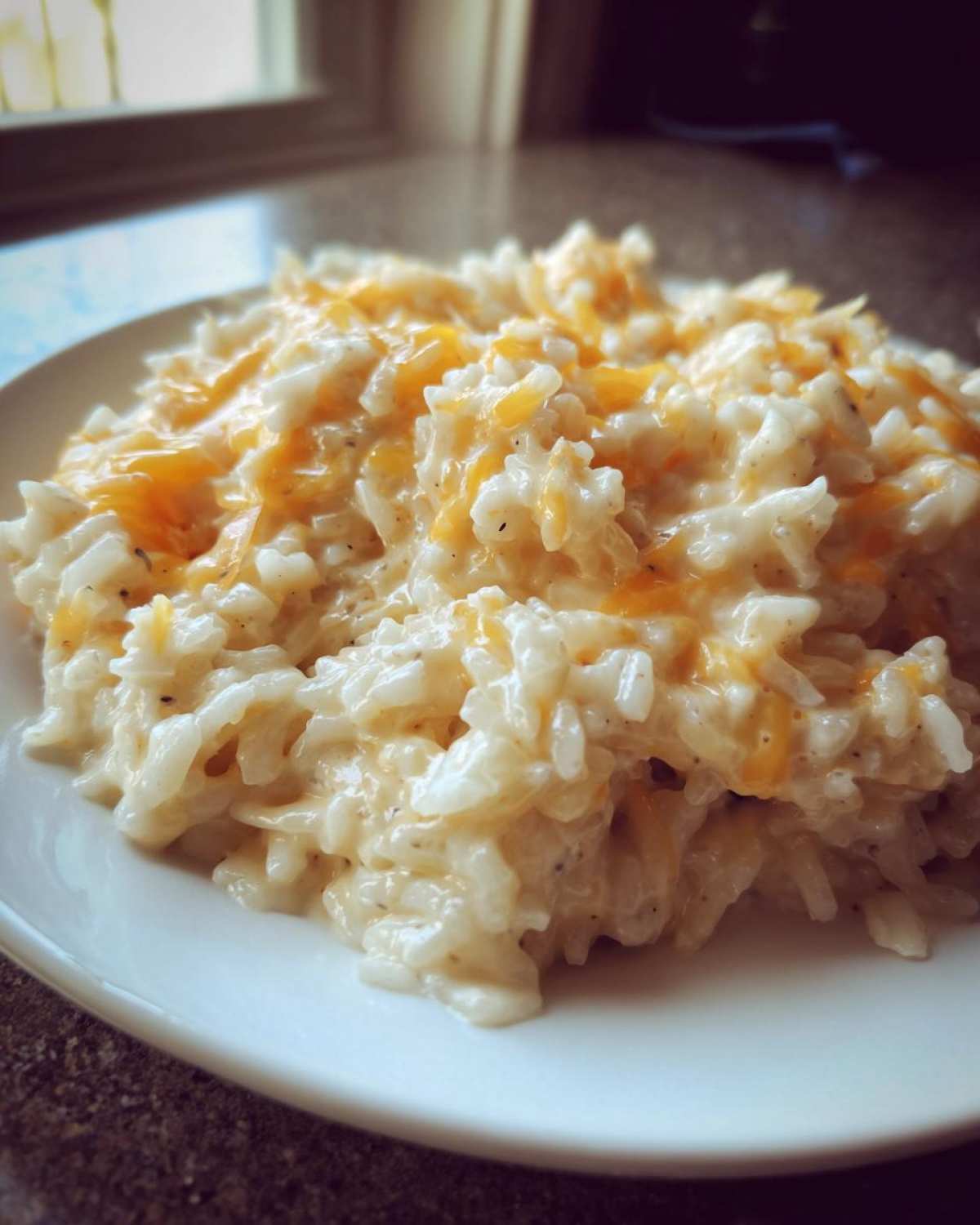 A close-up serving of creamy cheesy rice topped with melted shredded cheddar cheese on a white plate.