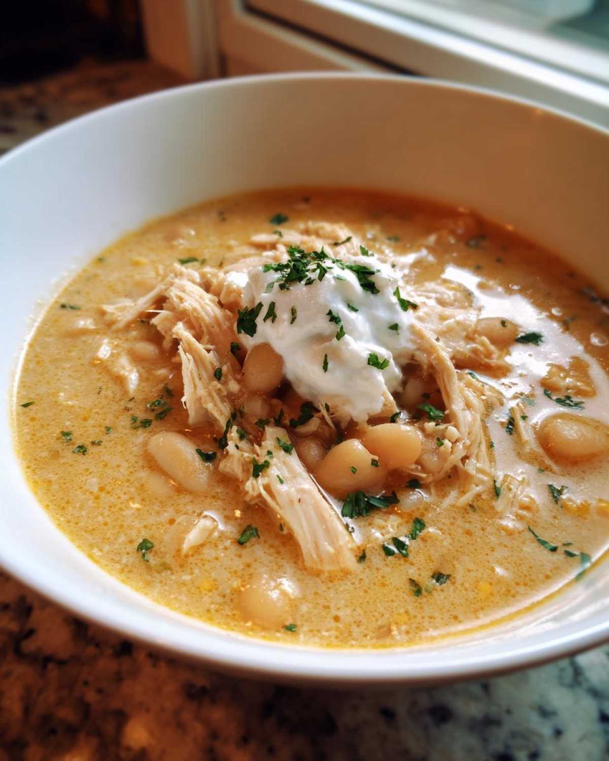 A close-up of a bowl of creamy crockpot white chicken chili topped with sour cream and parsley.