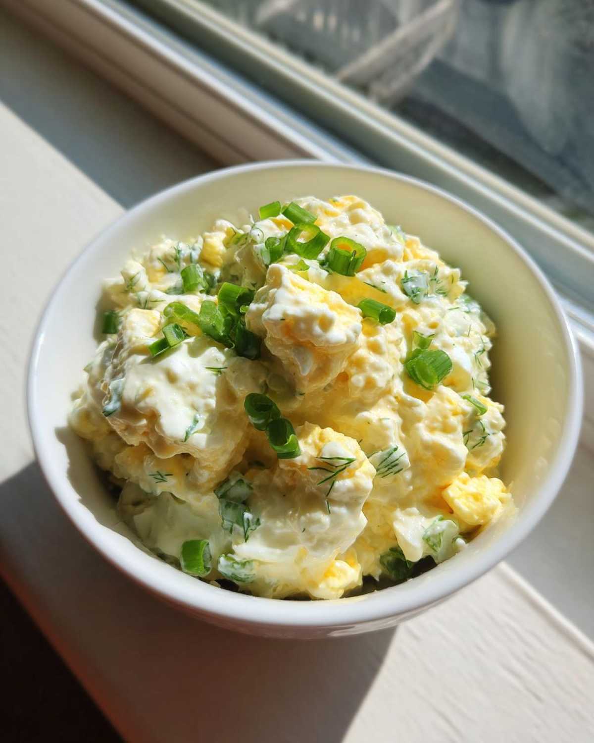 A close-up of creamy egg salad recipe served in a white bowl, topped with fresh chopped green onions.