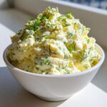 Close-up of a white bowl filled with creamy egg salad recipe, generously garnished with fresh green herbs.