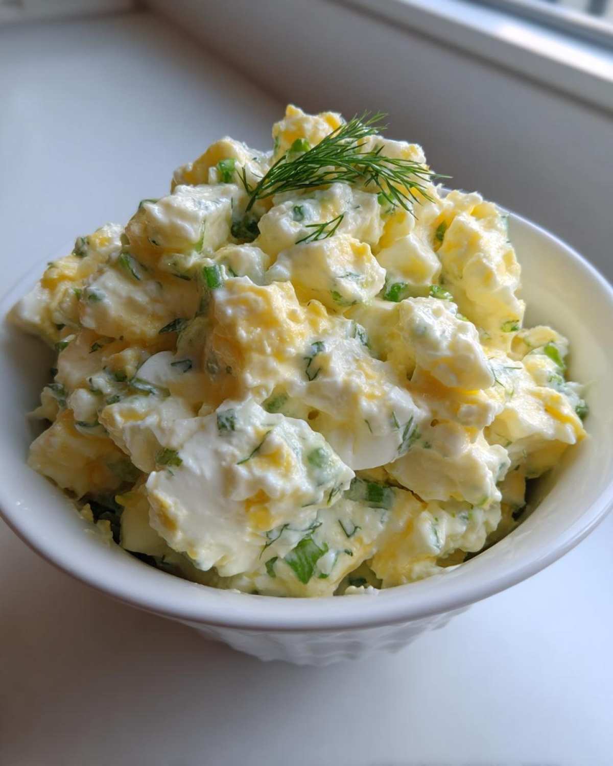 Close-up of creamy egg salad recipe, featuring chopped eggs, mayonnaise, and green herbs, garnished with fresh dill.