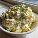 A bowl of creamy egg salad recipe, generously topped with fresh dill and chives, sitting by a sunny window.