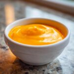 Close-up of smooth, bright orange nacho cheese sauce served in a small white dipping bowl.
