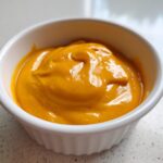 Close-up of rich, vibrant orange nacho cheese sauce served in a small white ramekin.