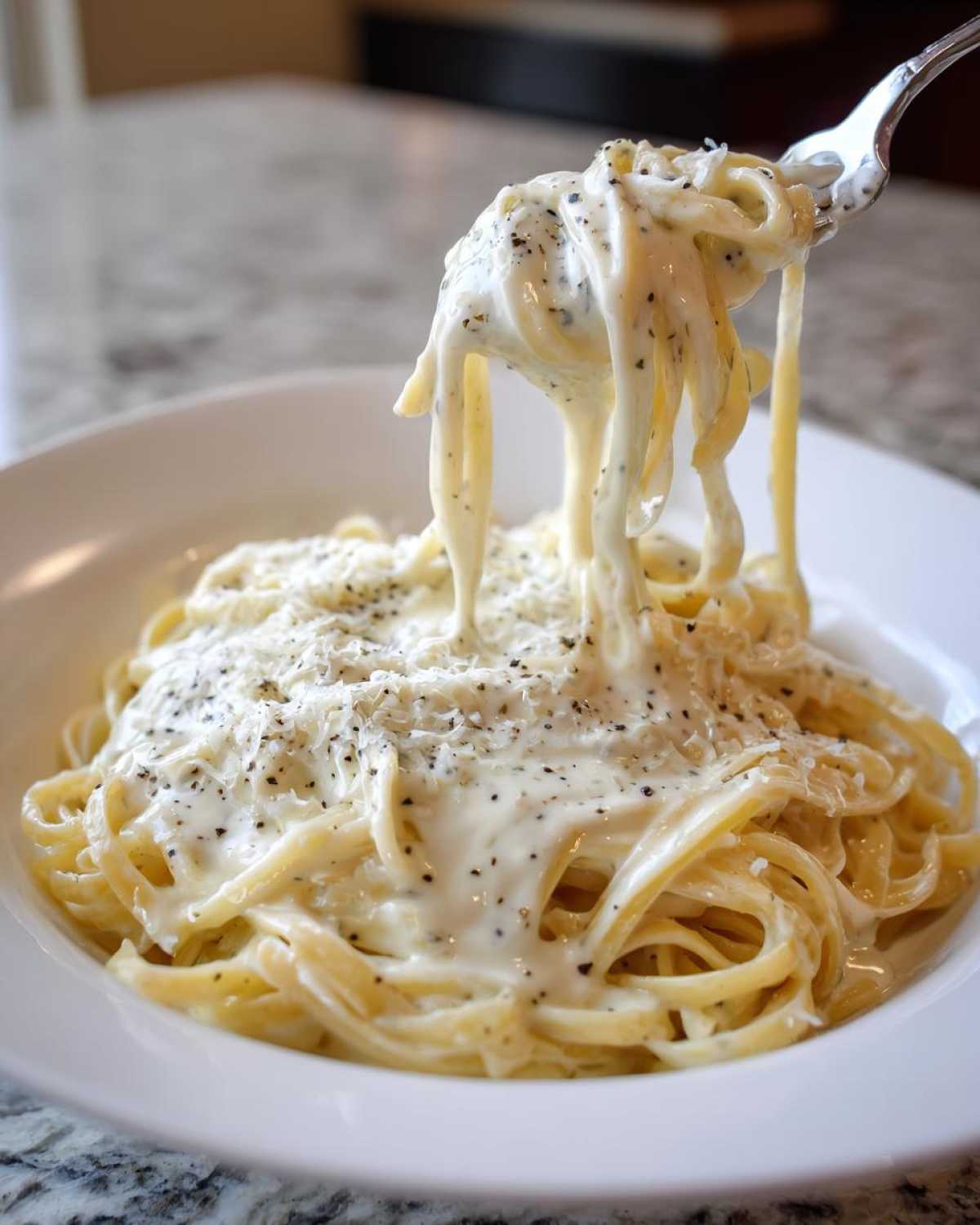 A fork lifting pasta coated in creamy Olive Garden Alfredo Sauce, topped with Parmesan and black pepper.