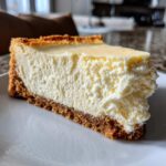Close-up of a thick slice of creamy Protein Cheesecake on a white plate showing the textured filling and graham cracker crust.
