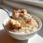 A spoonful of creamy rice pudding topped with cinnamon being lifted from a white bowl.