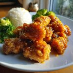 Close-up of crispy orange chicken recipe pieces coated in glaze and topped with zest, served with white rice and steamed broccoli.